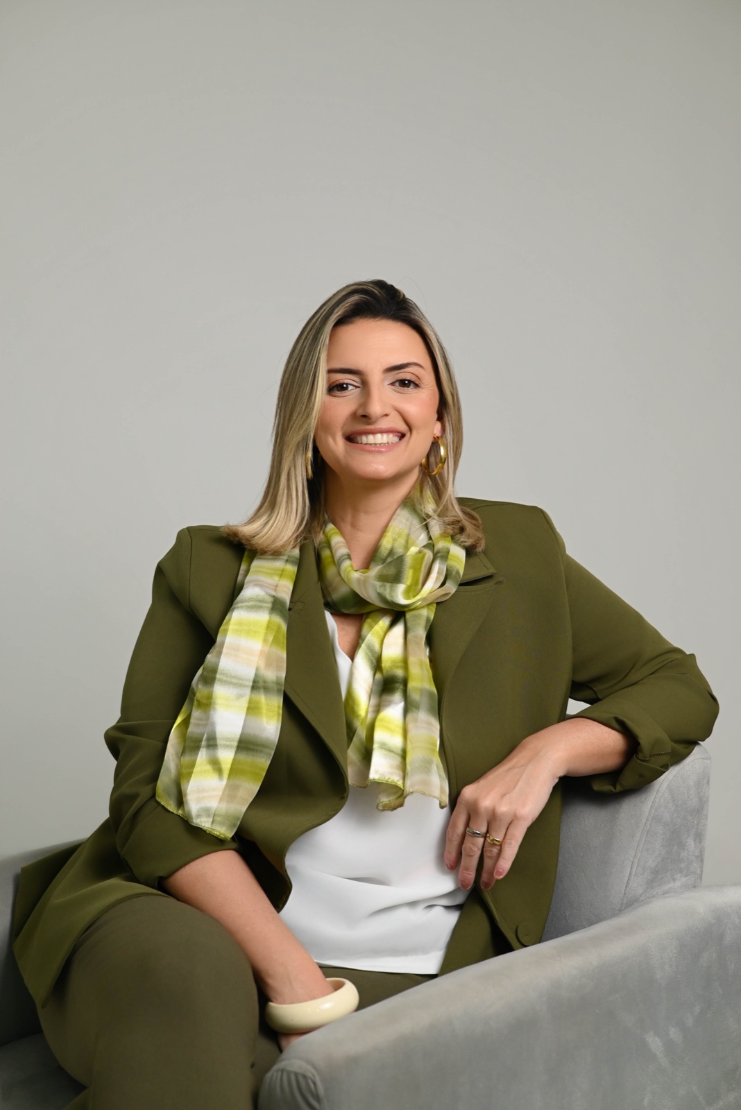 Luiza Palmerini rindo, sentada numa poltrona, de blazer verde e lenço